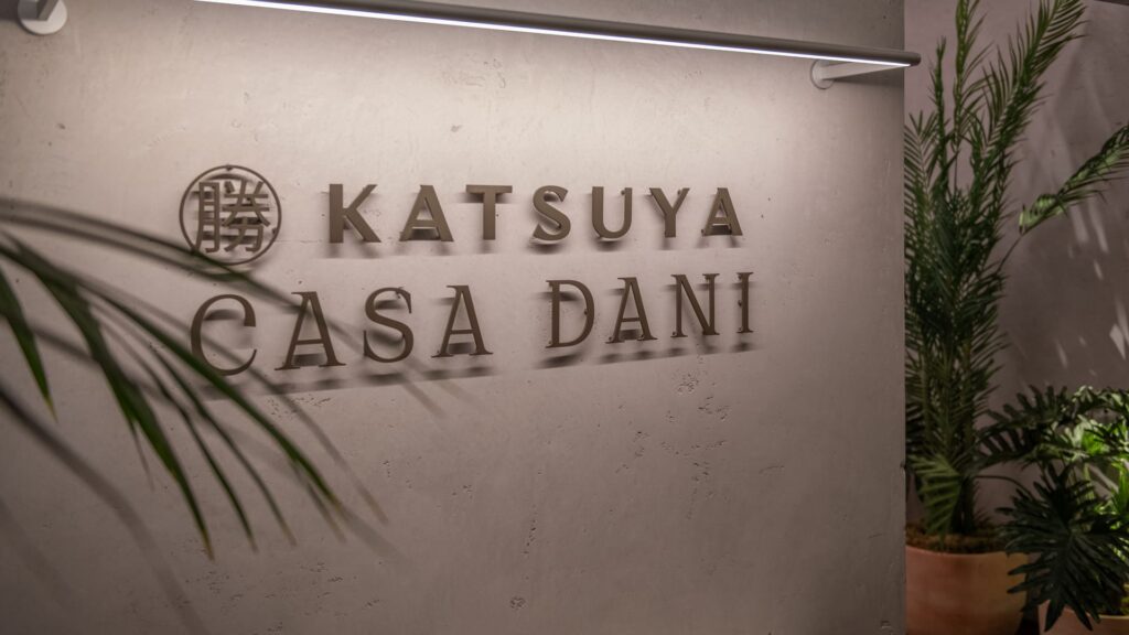 Wall signage for Katsuya and Casa Dani, with a modern brushed metal finish and soft lighting, surrounded by green plants.