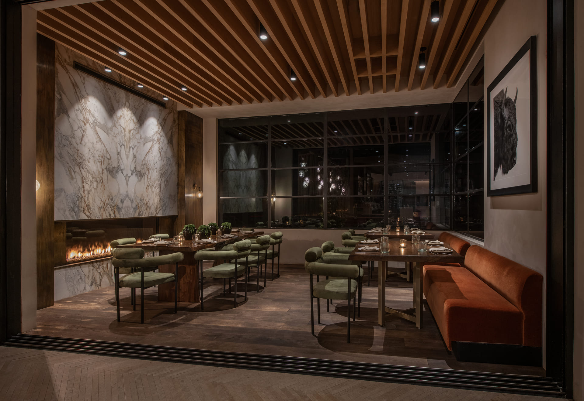 A stylish dining room with a long table, green cushioned chairs, and an orange velvet bench. The space features a marble accent wall with a fireplace, warm lighting, and wood panel ceilings, creating an elegant and cozy atmosphere.